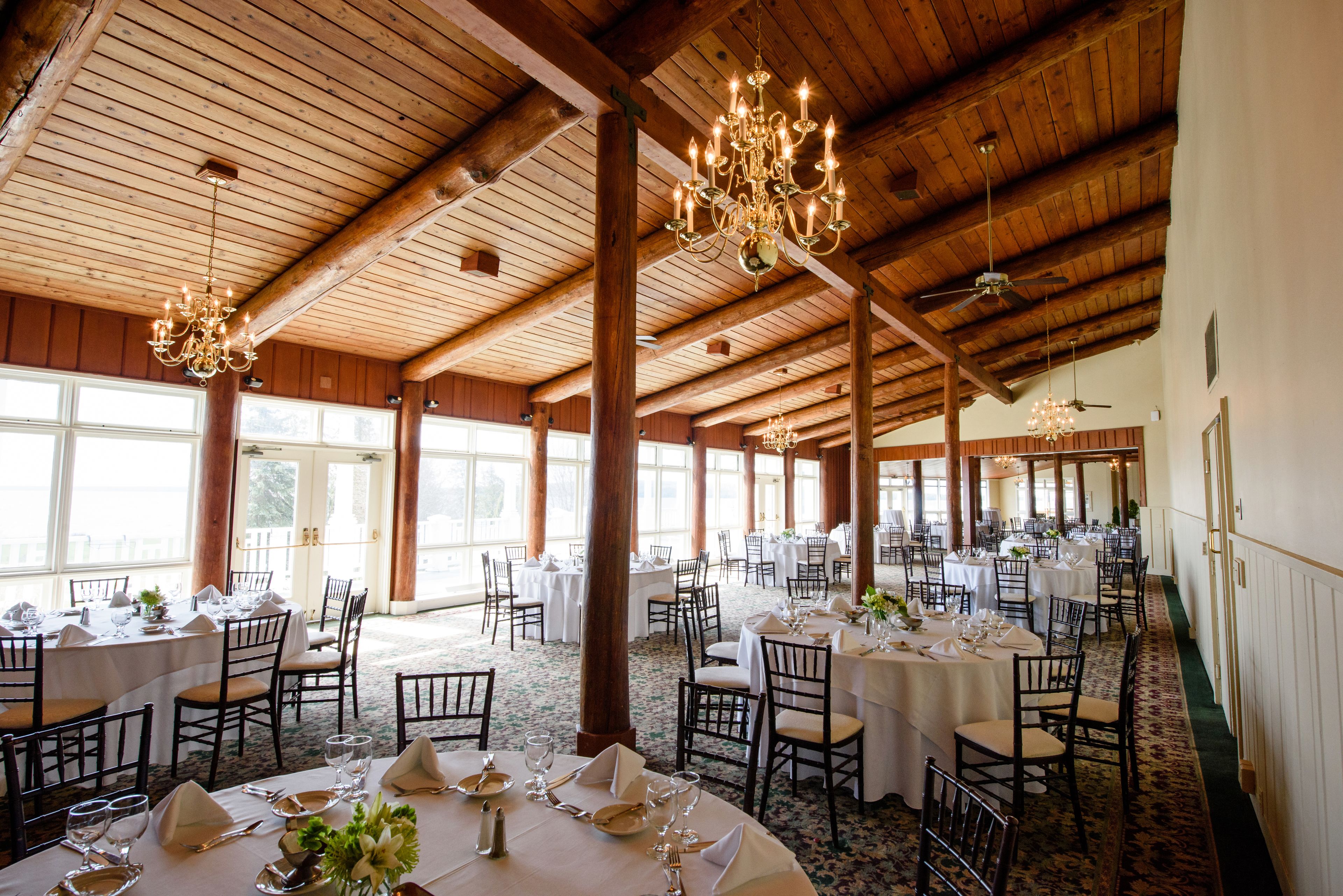 A banquet hall with round tables set for dining, wooden beams on the ceiling, and large windows allowing natural light.