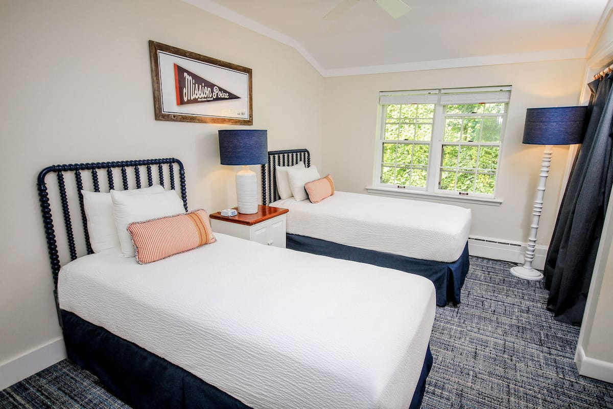 A hotel room with two beds, a window with a view of greenery, a framed sign on the wall, and a floor lamp.