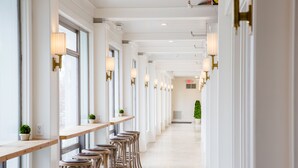 A long, narrow hallway with wooden tables and chairs, white walls, and wall-mounted lamps.