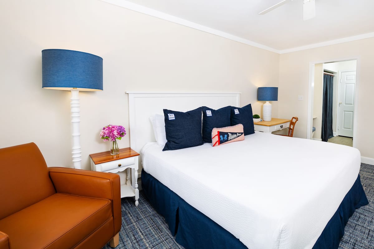 A hotel room with a large bed, a nightstand, a lamp, a chair, and a door leading to another room.
