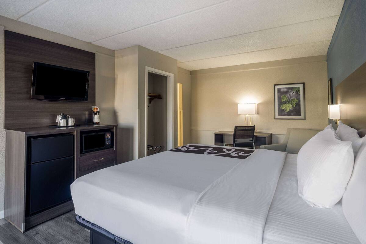 A hotel room with a bed, a TV mounted on the wall, a desk with a lamp, and a microwave.