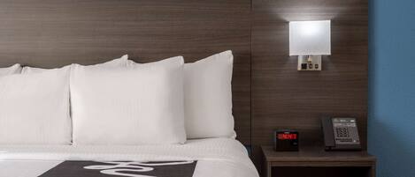 A hotel room with a large bed, white pillows, a dark headboard, a bedside table with a lamp, a digital clock, and a telephone.