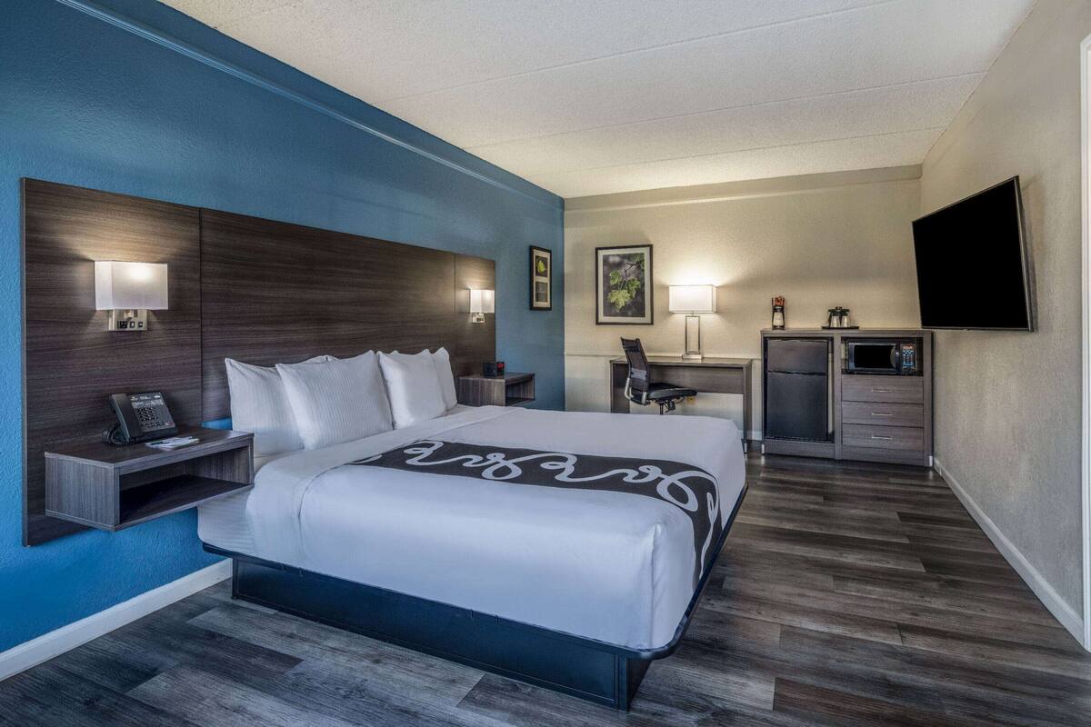 A hotel room with a bed, bedside table, lamp, desk, chair, and a flat-screen TV.