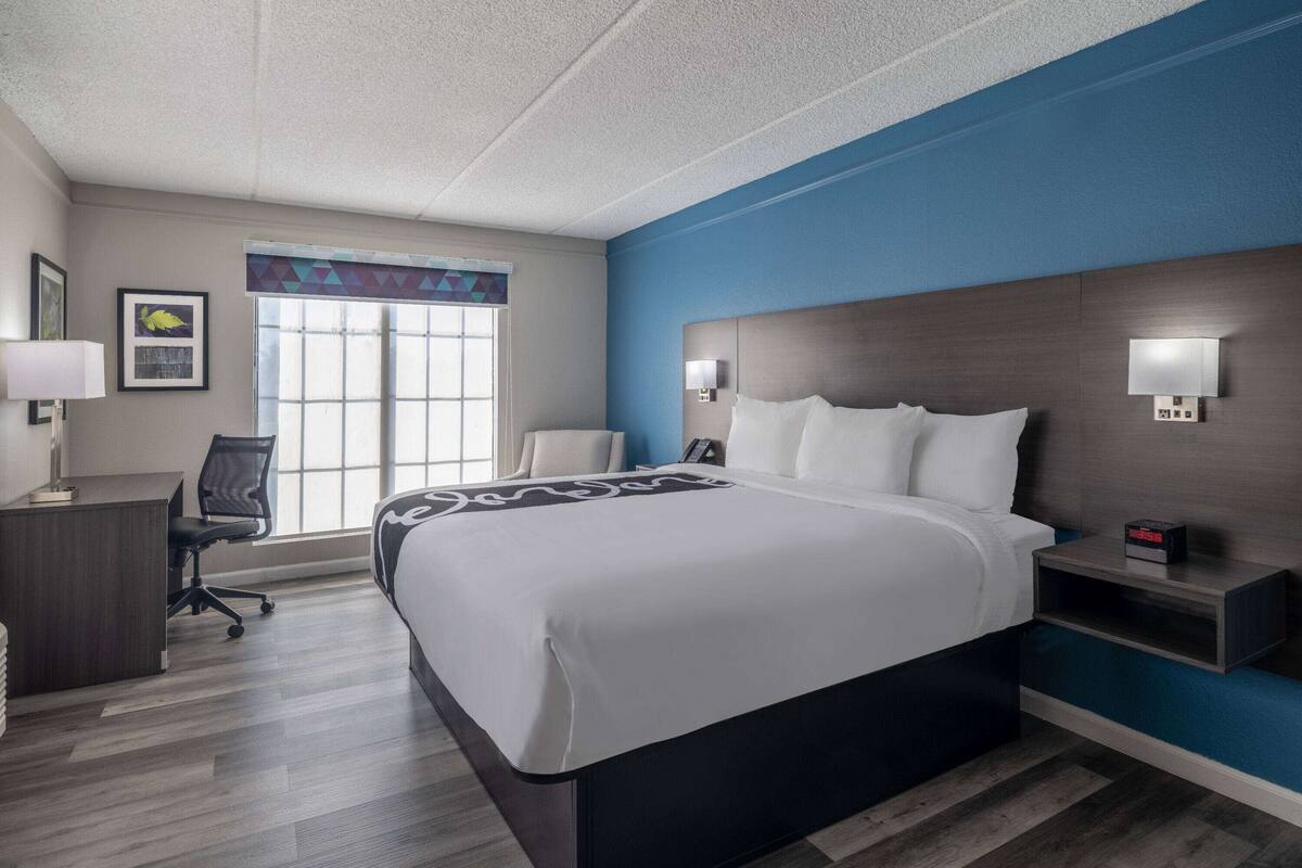 A hotel room with a large bed, a desk, and a chair. There is a window with a view of the outside.