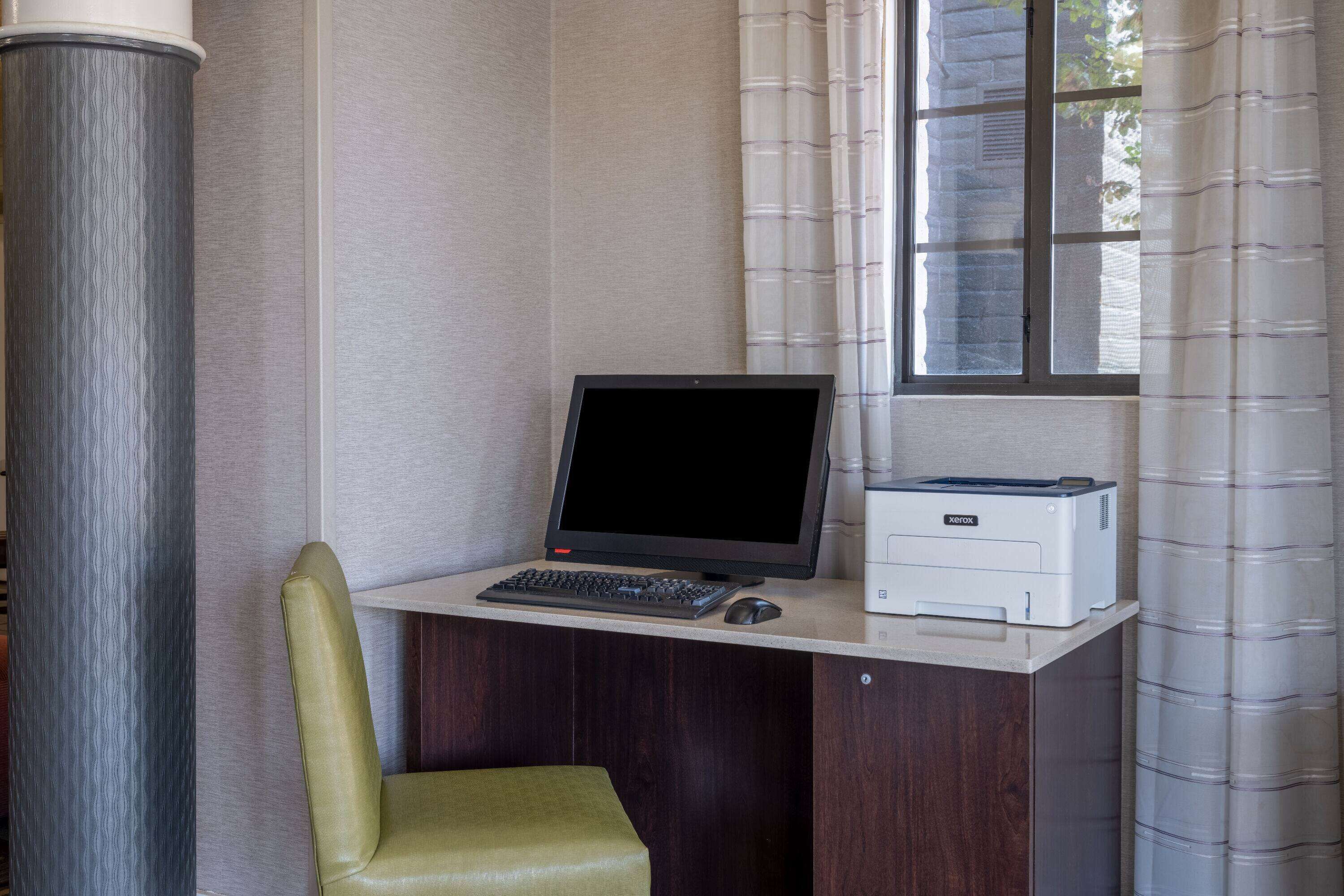 Ein Arbeitsplatz mit Computer, Tastatur, Maus und Drucker auf einem Schreibtisch, einem grünen Stuhl und einem Fenster mit Vorhängen.
