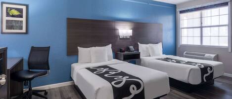 A hotel room with two beds, a desk, a chair, a nightstand, and a window.