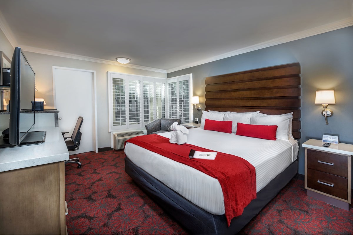 A hotel room with a large bed, a desk with a computer, a chair, a nightstand, and a window with blinds.