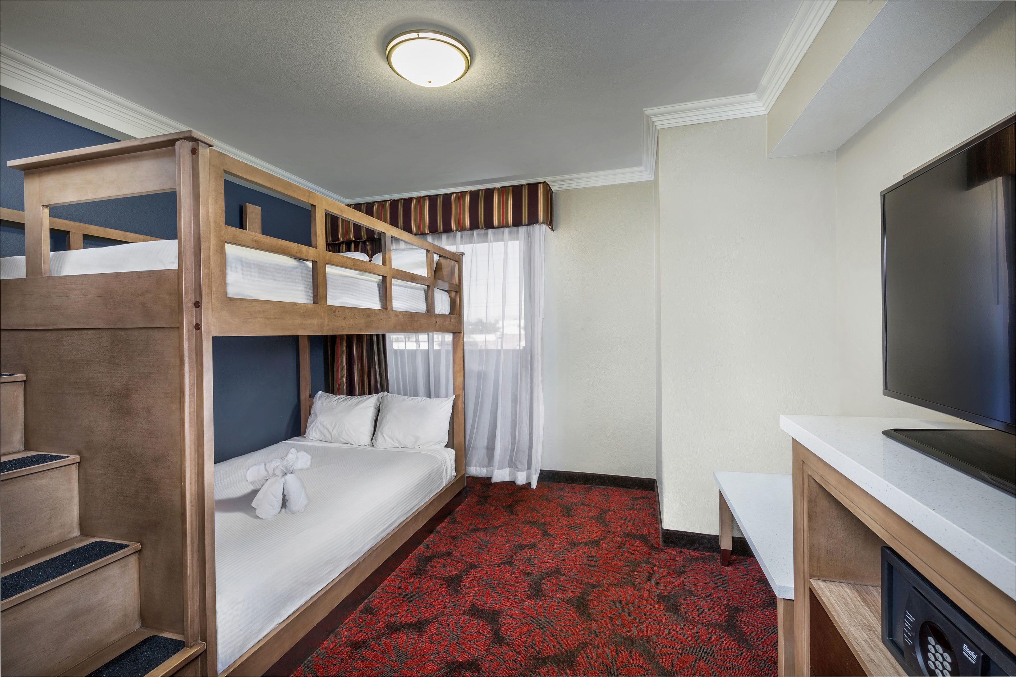 A hotel room with a bunk bed, a desk with a flat-screen TV, a red patterned carpet, and a window with sheer curtains.