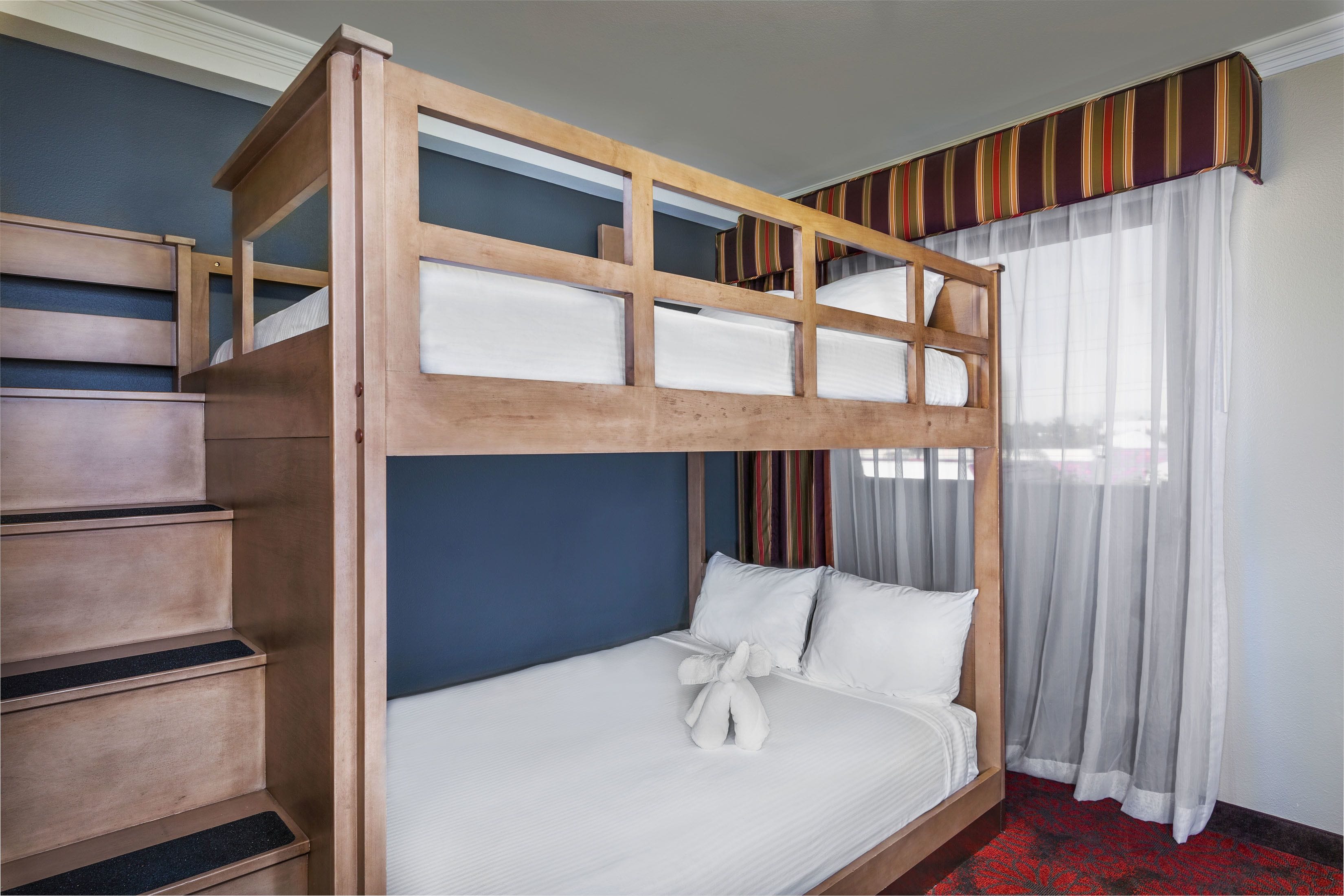 Un lit superposé avec une structure en bois et du linge de lit blanc, une commode en bois et une fenêtre avec des rideaux légers.