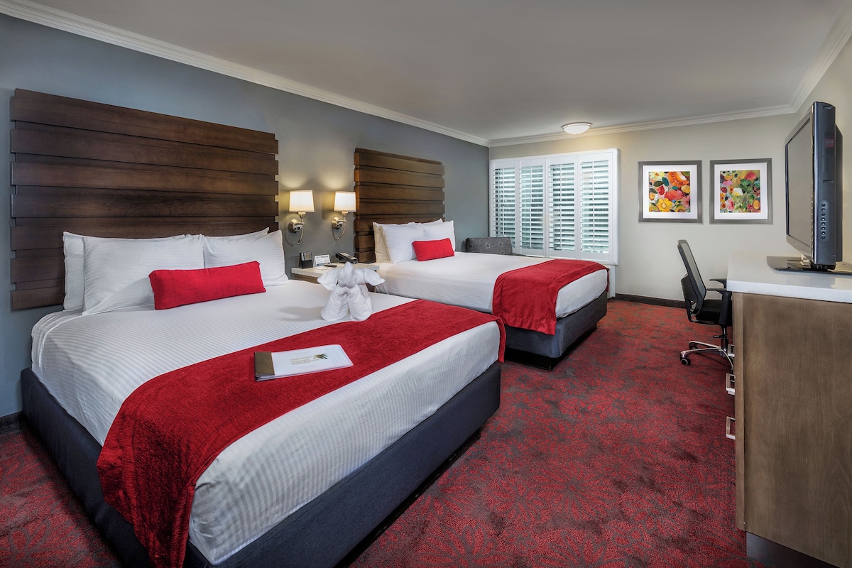 A hotel room with two beds, a desk, a chair, and artwork on the wall.
