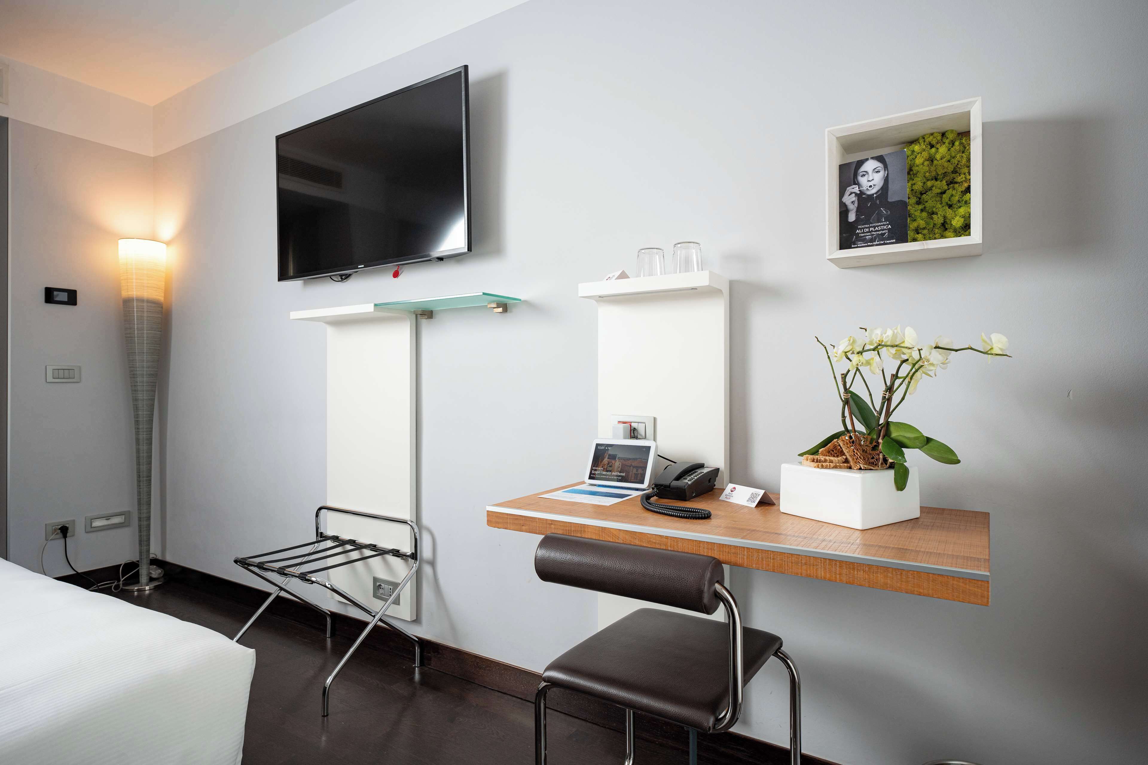 Ein Hotelzimmer mit einem Bett, einem Schreibtisch mit Laptop, einem Sessel, einem Fernseher, einem Wandregal mit Bilderrahmen und einer Topfpflanze.