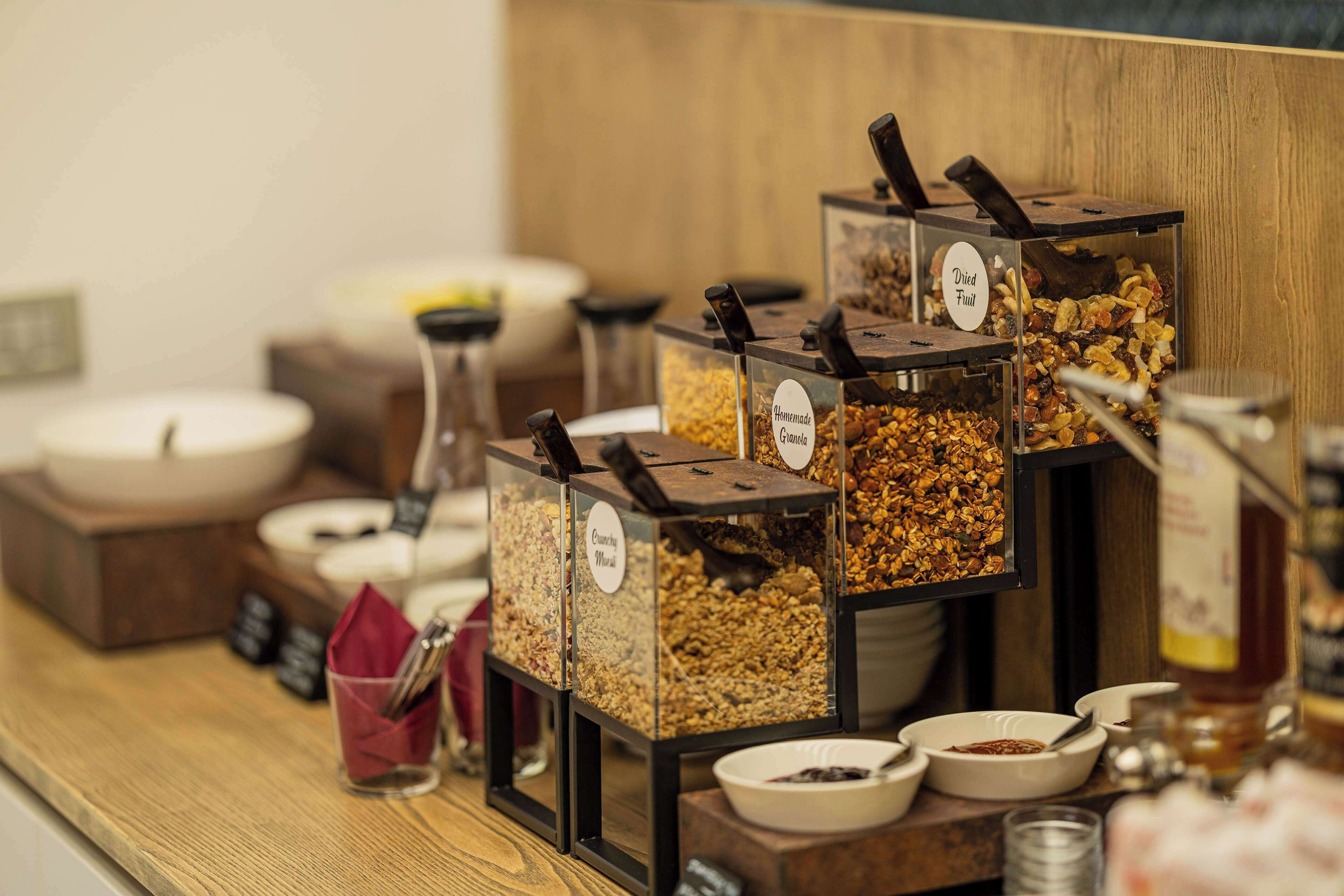 Una barra de desayuno con recipientes etiquetados de granola, fruta seca y otros cereales.
