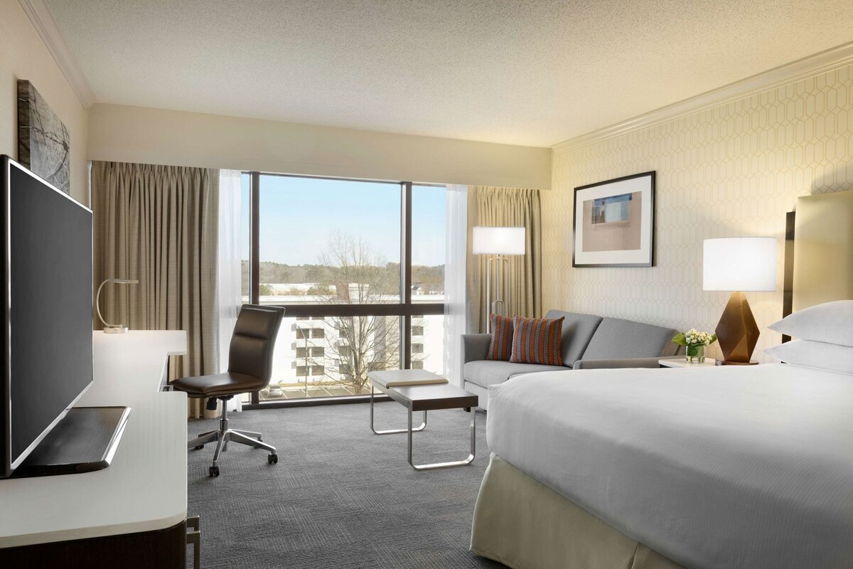 A hotel room with a bed, a desk with a computer, a chair, a small table, a sofa, and a large window offering a view.