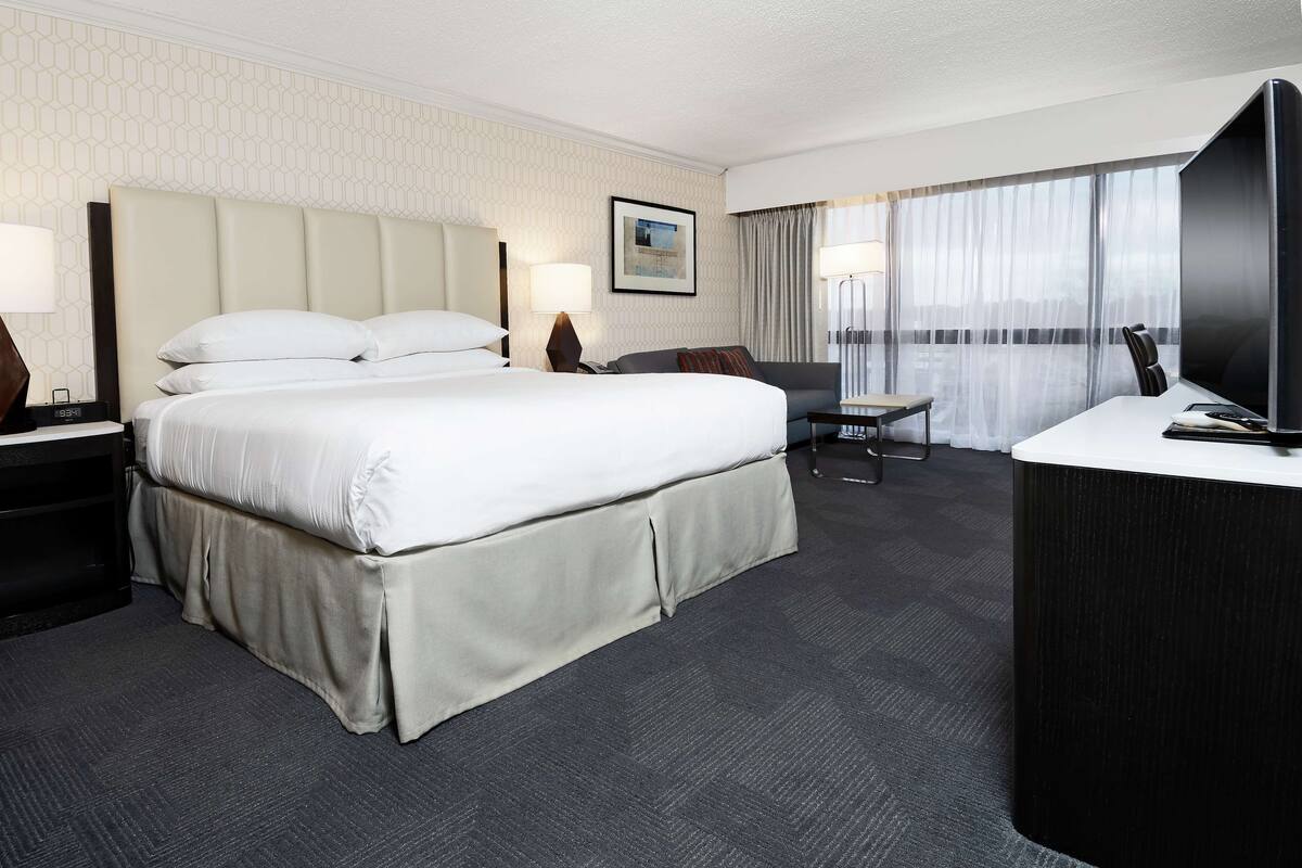 A hotel room with a large bed, a desk with a TV, a chair, and a desk lamp.