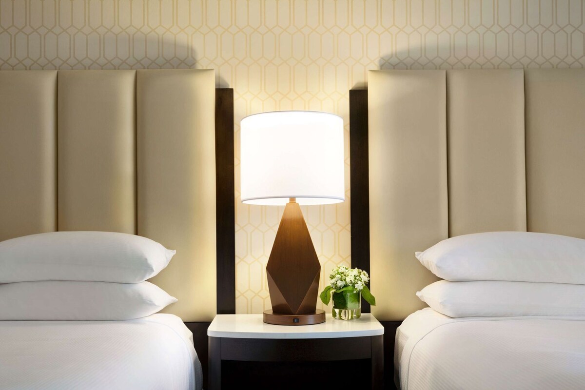 A hotel room with two beds, a nightstand with a lamp, and a vase with flowers.