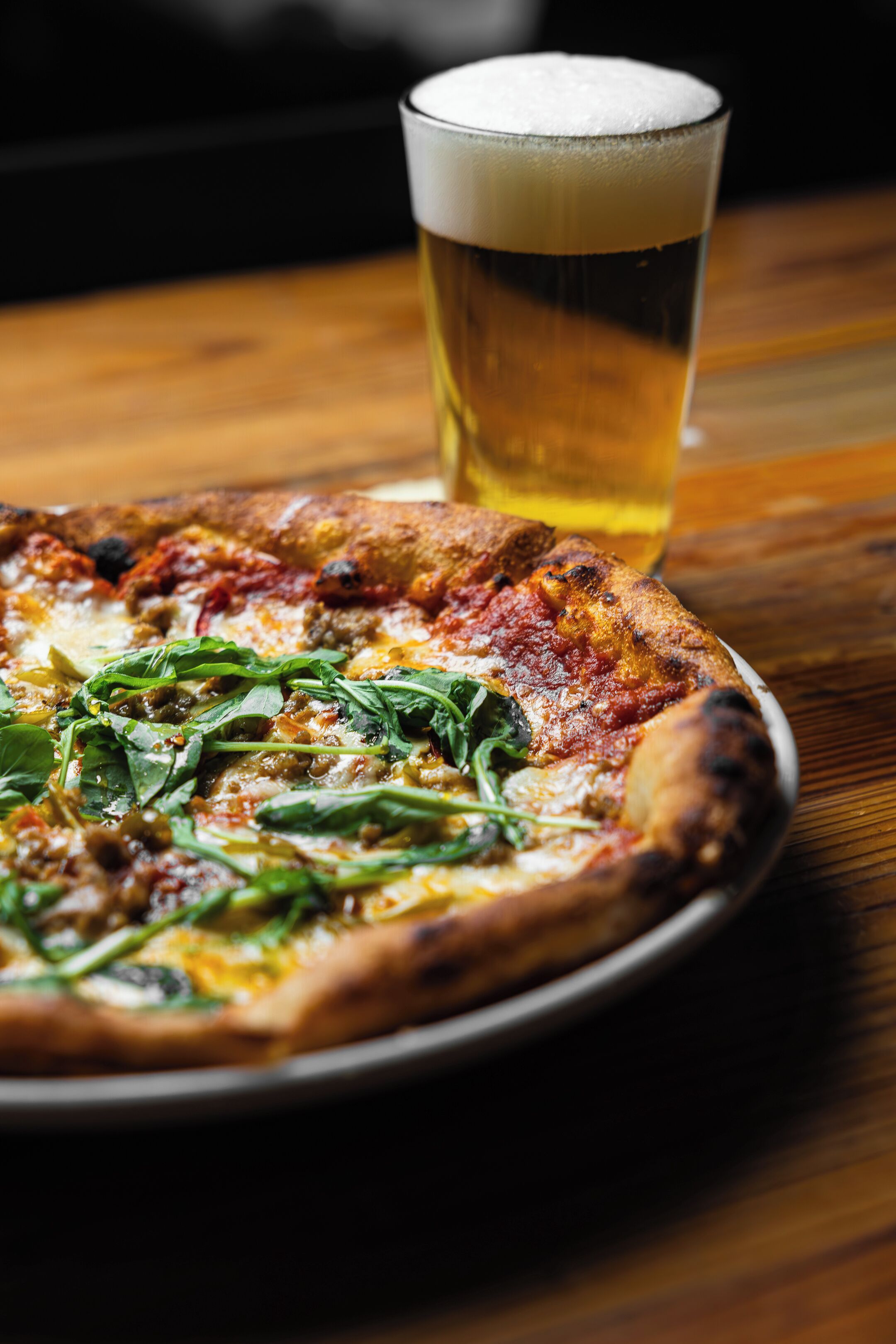 Pizza con espinacas sobre un plato, una jarra de cerveza y una mesa de madera.
