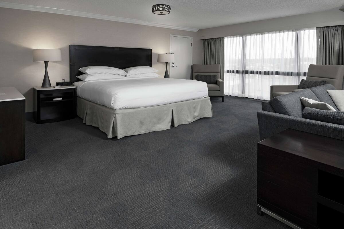 A hotel room with a large bed, two bedside tables with lamps, a dark wood dresser, a grey sofa, and a large window with sheer curtains.