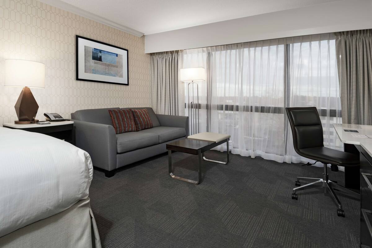 A hotel room with a bed, a sofa, a small table, a desk with a chair, and a large window with curtains.