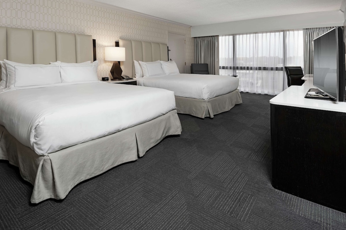 A hotel room with two beds, a desk, a TV, and a window with curtains.