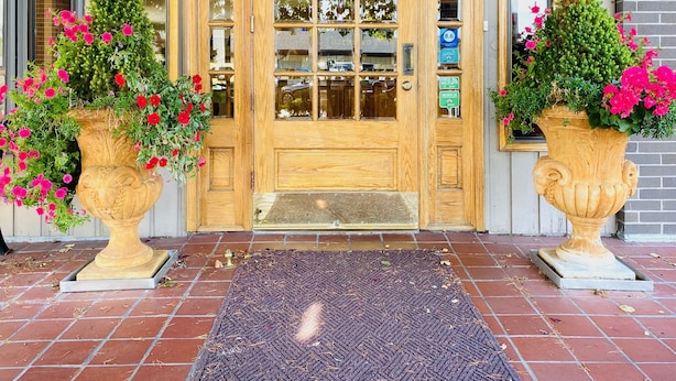 A wooden front door with glass panels, flanked by two large flower pots with pink flowers, and a dark woven mat at the entrance. - Coachman Inn & Suites (Oak Harbor)