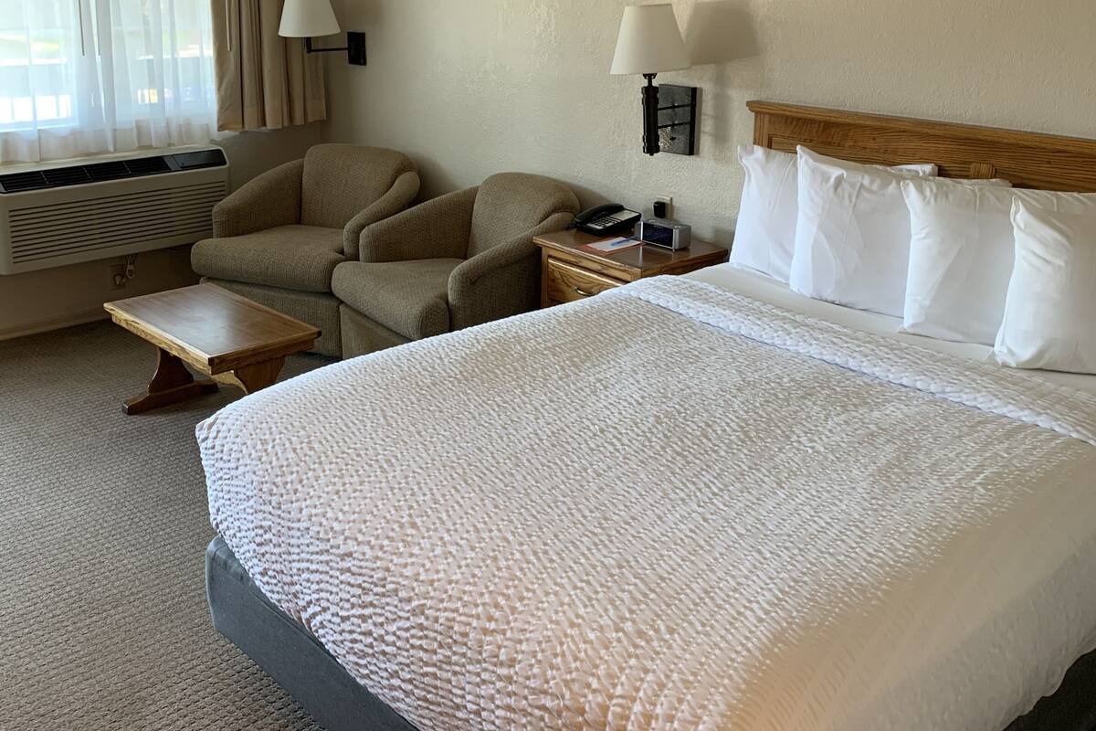 A hotel room with a large bed, two armchairs, a small table, a bedside lamp, and a window with curtains.