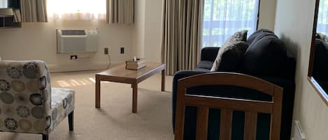 Una sala de estar con un sofá, un sillón y una mesa de centro de madera. Hay un televisor de pantalla plana montado en la pared, y un equipo de aire acondicionado también en la pared.