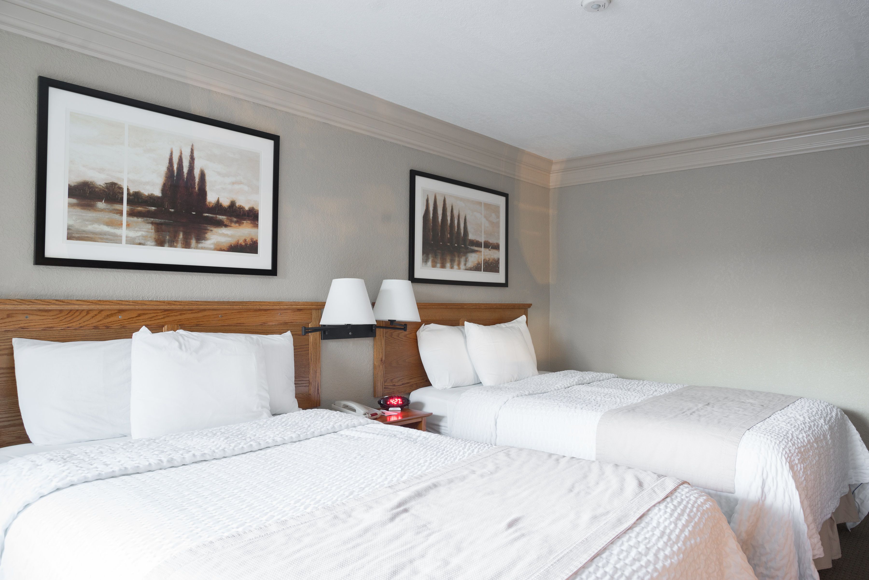 Ein Hotelzimmer mit zwei Betten, weißer Bettwäsche, einem Holzkopfteil, einem Nachttisch mit Lampe sowie zwei gerahmten Bildern an der Wand.