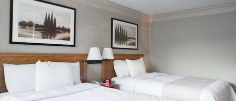Ein Hotelzimmer mit zwei Betten, weißer Bettwäsche, einem Holzkopfteil, einem Nachttisch mit Lampe sowie zwei gerahmten Bildern an der Wand.