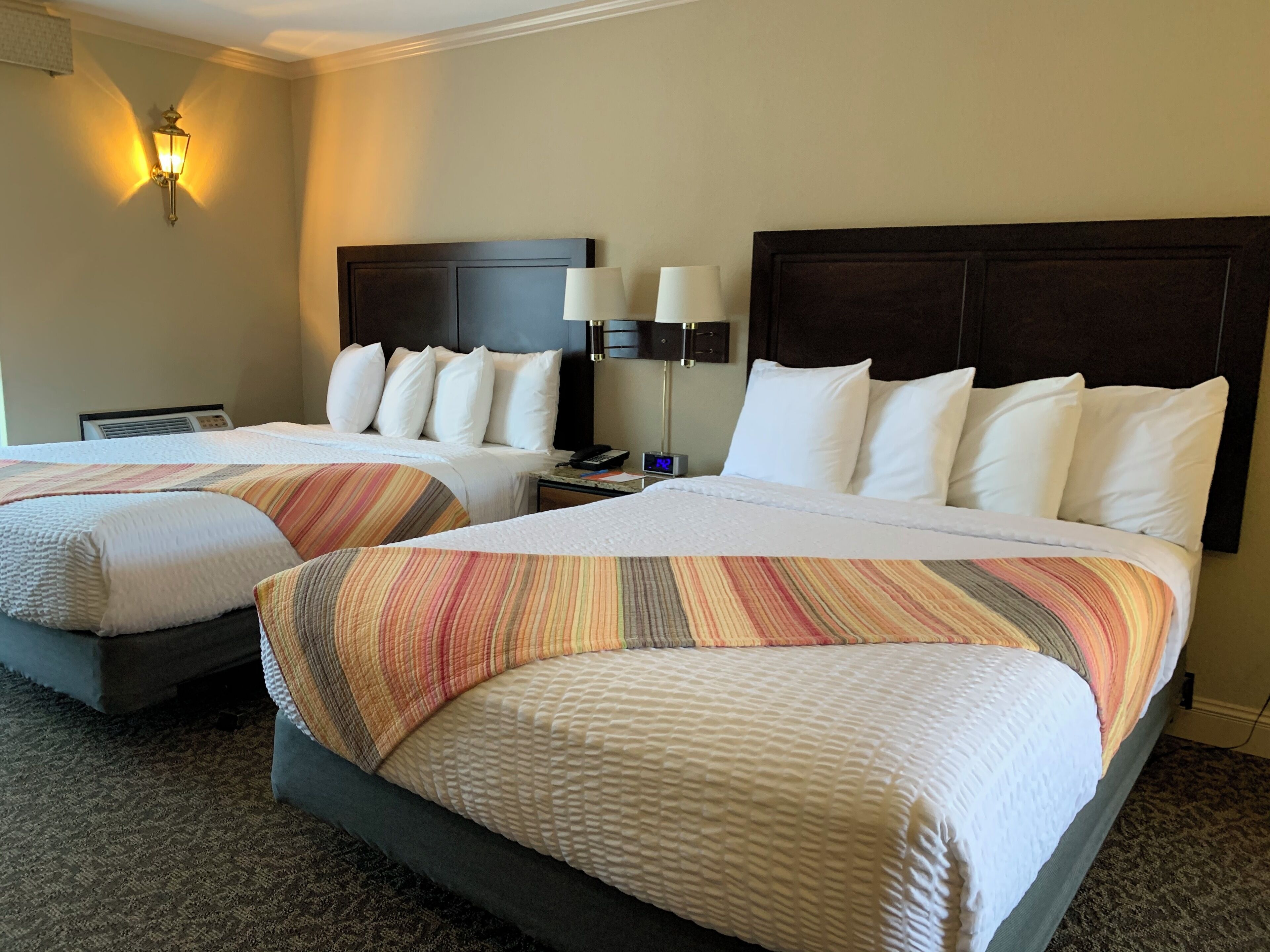 Two double beds with striped blankets, a dark headboard, a bedside table with a lamp, and a wall-mounted light fixture.
