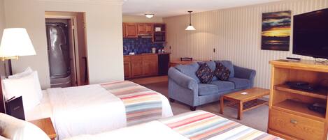 A hotel room with two beds, a sofa, a TV, and a kitchen area in the background.