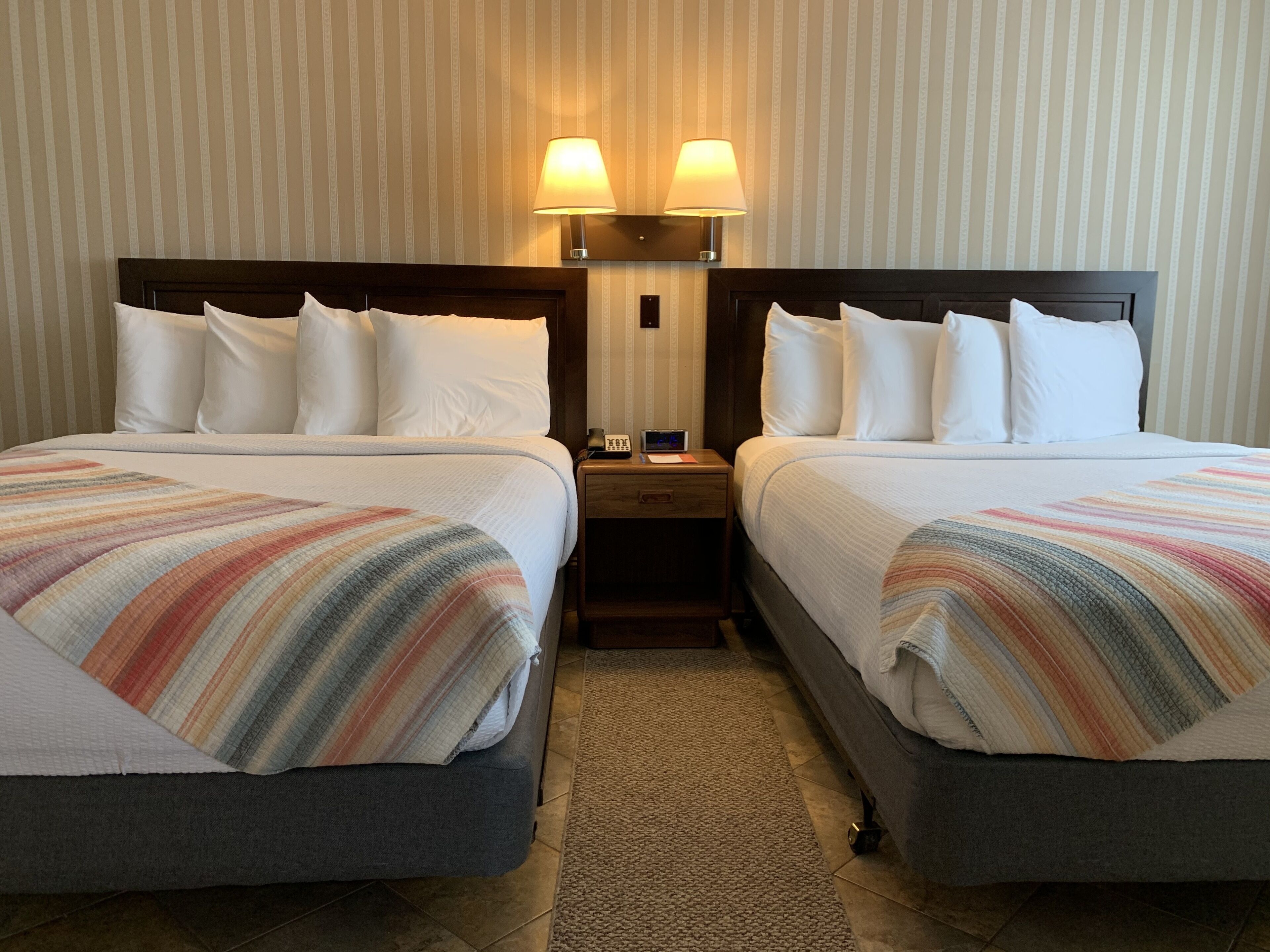Quarto de hotel com duas camas, cada uma com lençóis brancos e cobertores listrados. Há duas luminárias de cabeceira e uma mesinha de cabeceira com um telefone e uma pequena mesa.