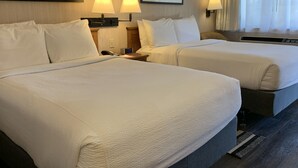 A hotel room with two beds, each with white linens, a bedside table with a lamp, and a clock. There are framed pictures on the wall and a window with curtains.