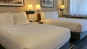 A hotel room with two beds, each with white linens, a bedside table with a lamp, and a clock. There are framed pictures on the wall and a window with curtains.