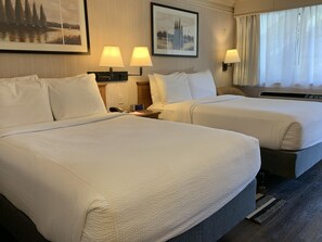 A hotel room with two beds, each with white linens, a bedside table with a lamp, and a clock. There are framed pictures on the wall and a window with curtains.