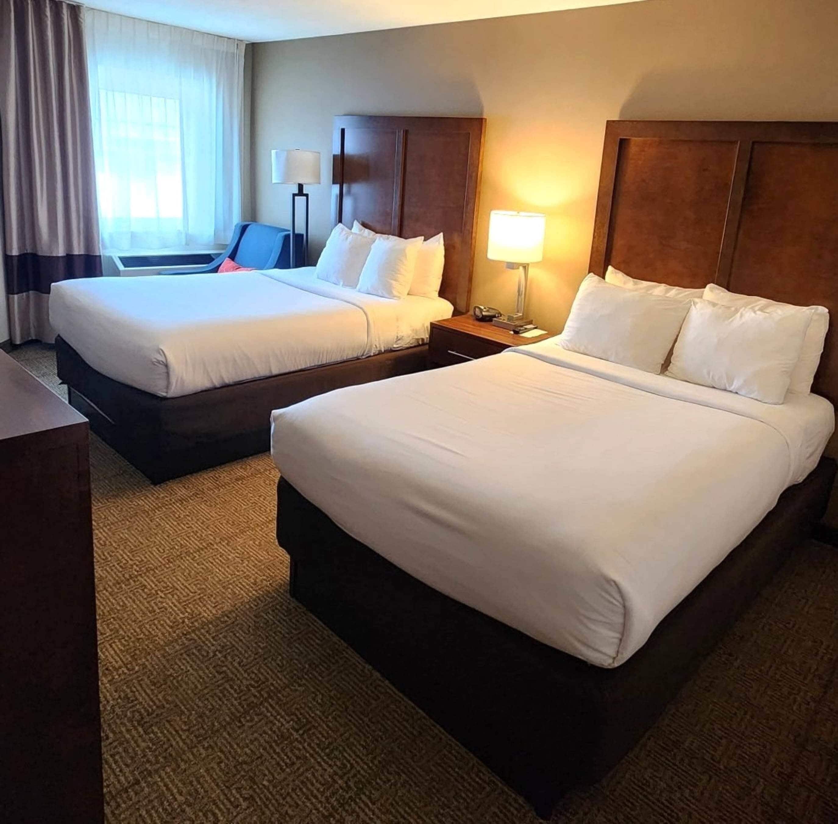 A hotel room with two beds, a desk, a chair, a lamp, and a window with curtains.