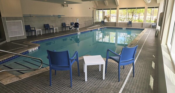 An indoor swimming pool with blue chairs and a white table, surrounded by windows.