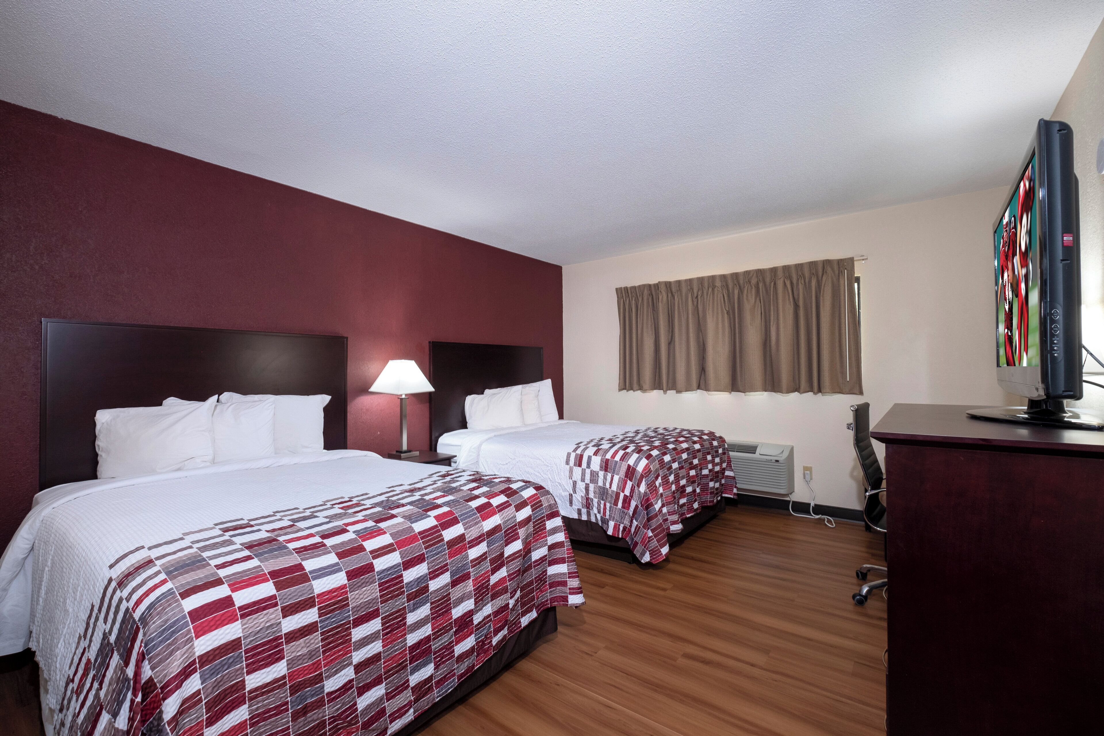 A hotel room with two beds, a desk, a television, and a window with curtains.
