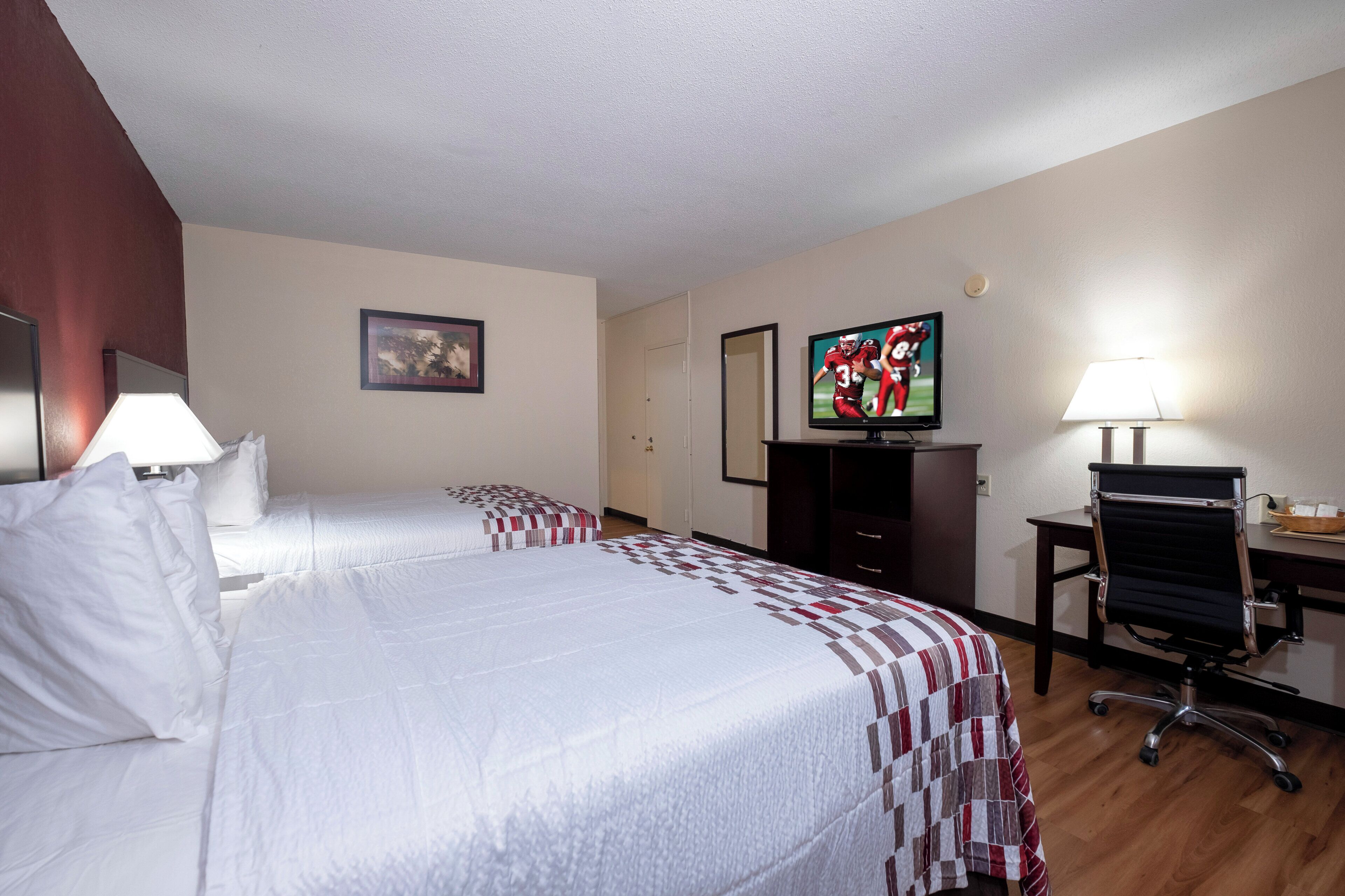 A hotel room with two beds, a desk with a chair, a television, and a lamp.