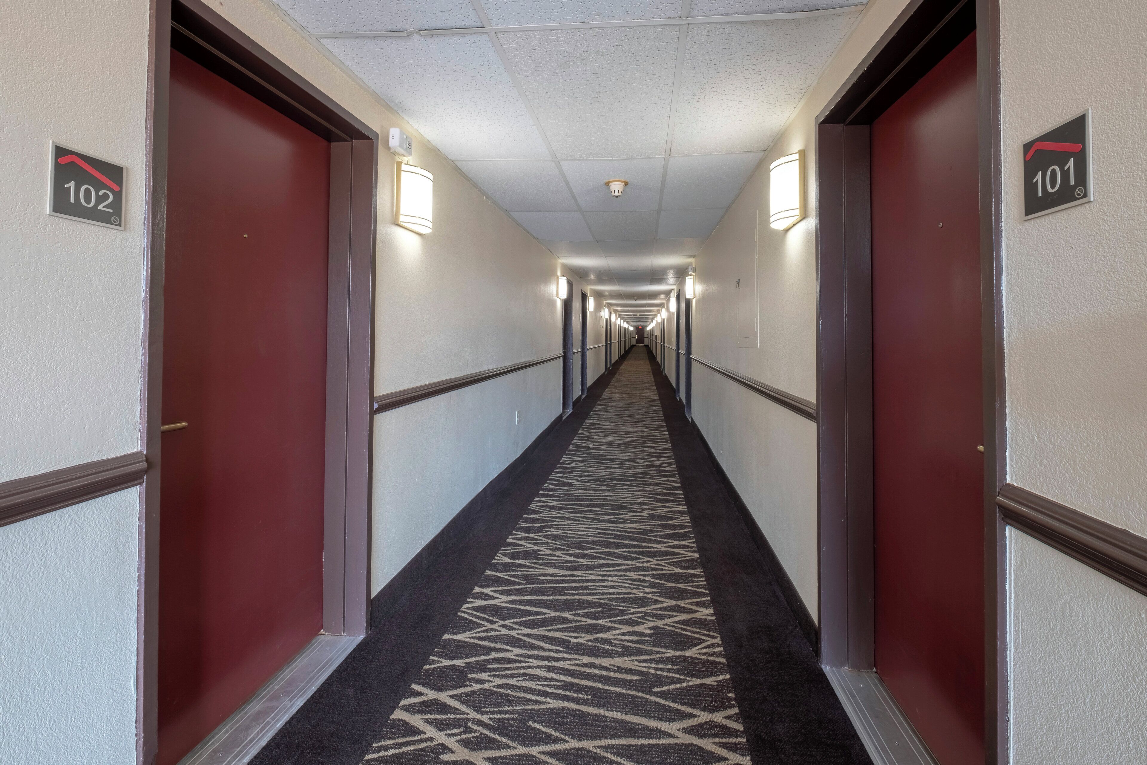 Een hotelgang met deuren genummerd 101 en 102, bekleed met een geometrisch patroon, en voorzien van wandlampen.