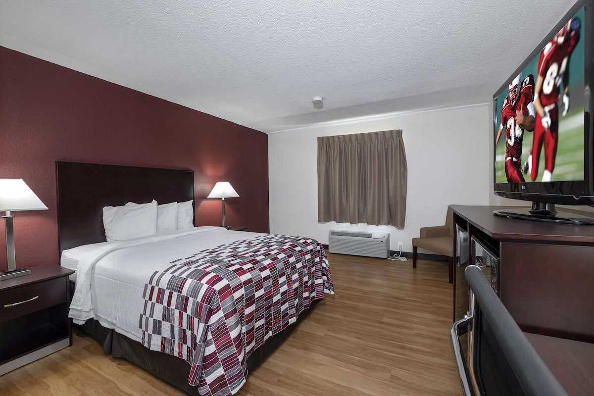 A hotel room with a large bed, two bedside tables with lamps, a flat-screen TV, a desk with a chair, and a window with curtains.