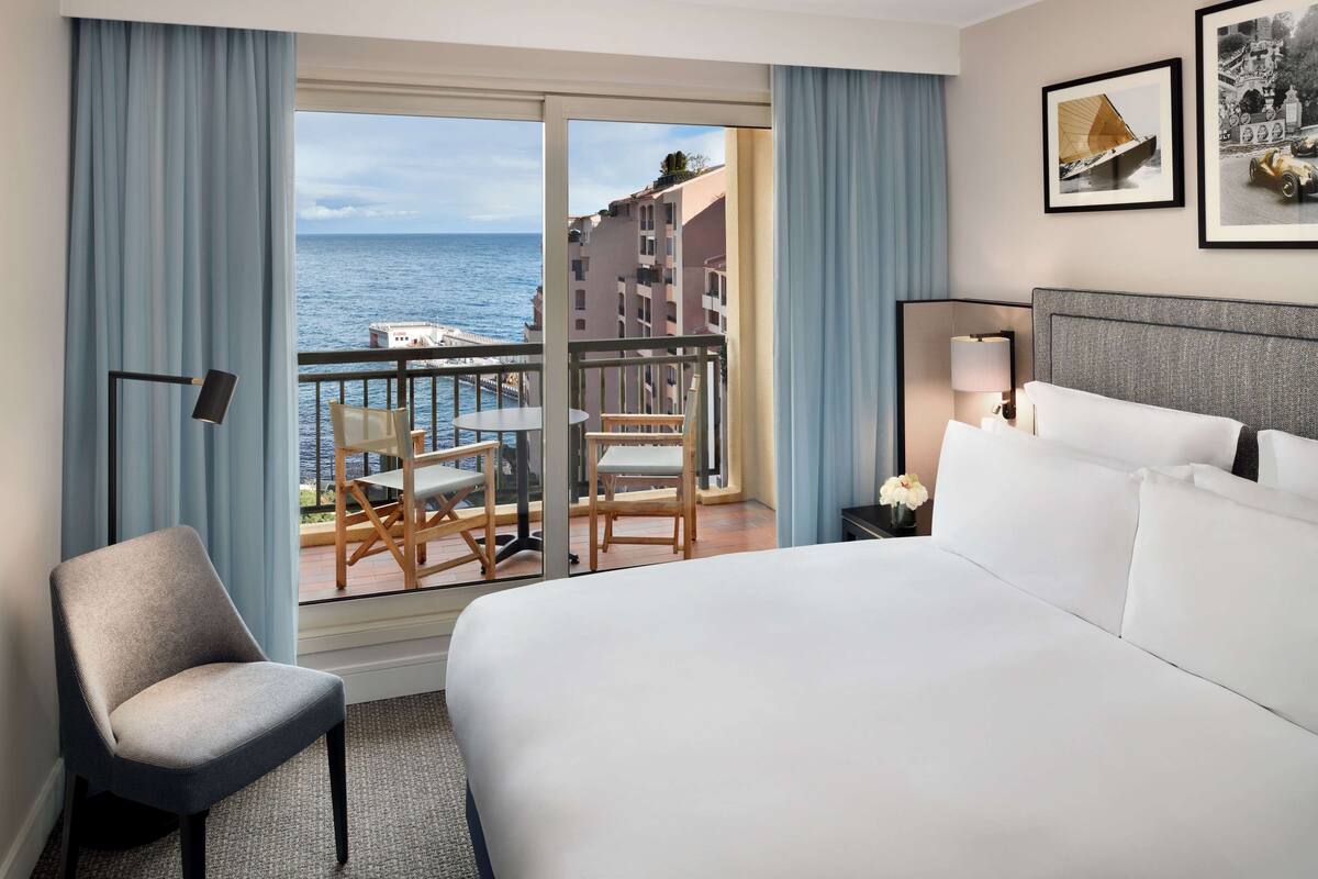 Ett hotellrum med en stor säng, en stol, ett bord med stolar och en balkong med utsikt över havet och byggnaderna.