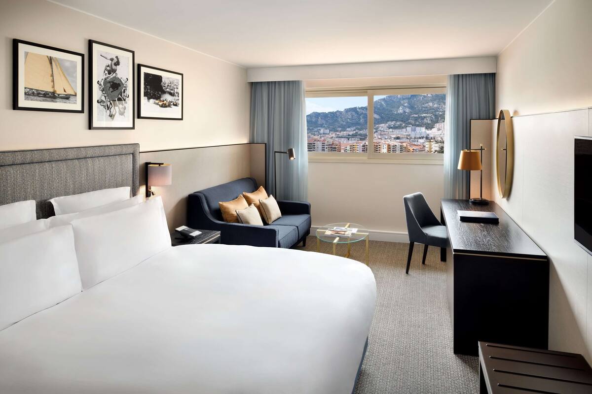 Ett hotellrum med en stor säng, en soffa, ett skrivbord och en stol. På väggen hänger inramade tavlor och genom fönstret har man utsikt över staden.