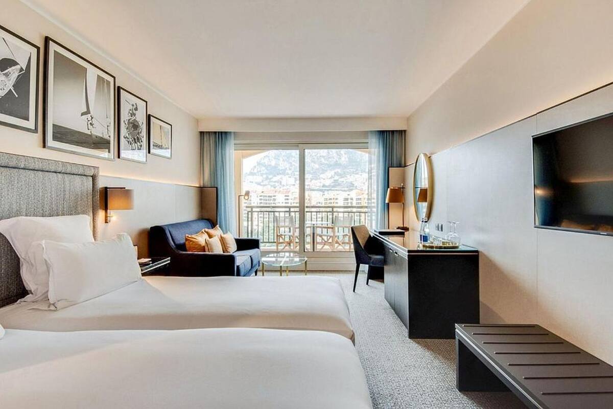 Ett hotellrum med två sängar, ett skrivbord, en stol, en TV och en balkong med utsikt över byggnader.