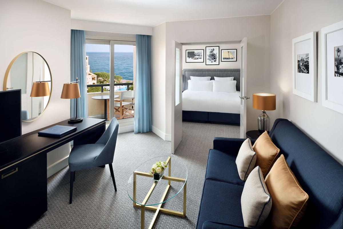 Ett hotellrum med ett skrivbord, en stol och ett soffbord i glas med blommor. Det finns en säng med utsikt över havet och en balkong med ett bord och stolar.
