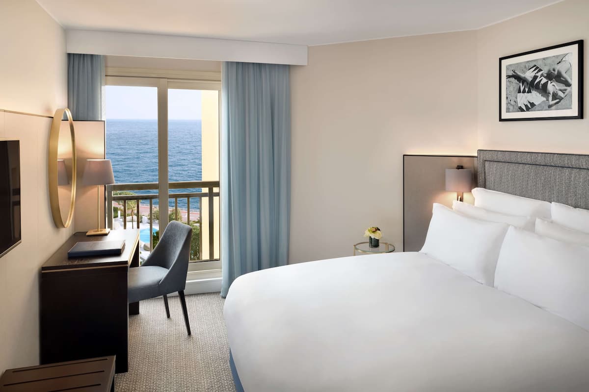 Ett hotellrum med en stor säng, ett skrivbord med en lampa, en stol och utsikt över havet genom ett stort fönster.