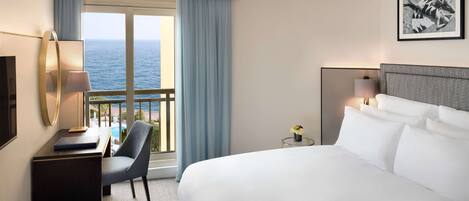 Une chambre d’hôtel avec un grand lit, un bureau avec une lampe, une chaise et une vue sur l’océan par une grande fenêtre.