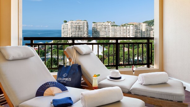 A balcony with two lounge chairs, a fan, a bag, a hat, and a glass of drink. - Columbus Hotel Monte-Carlo, Curio Collection by Hilton (Monaco)