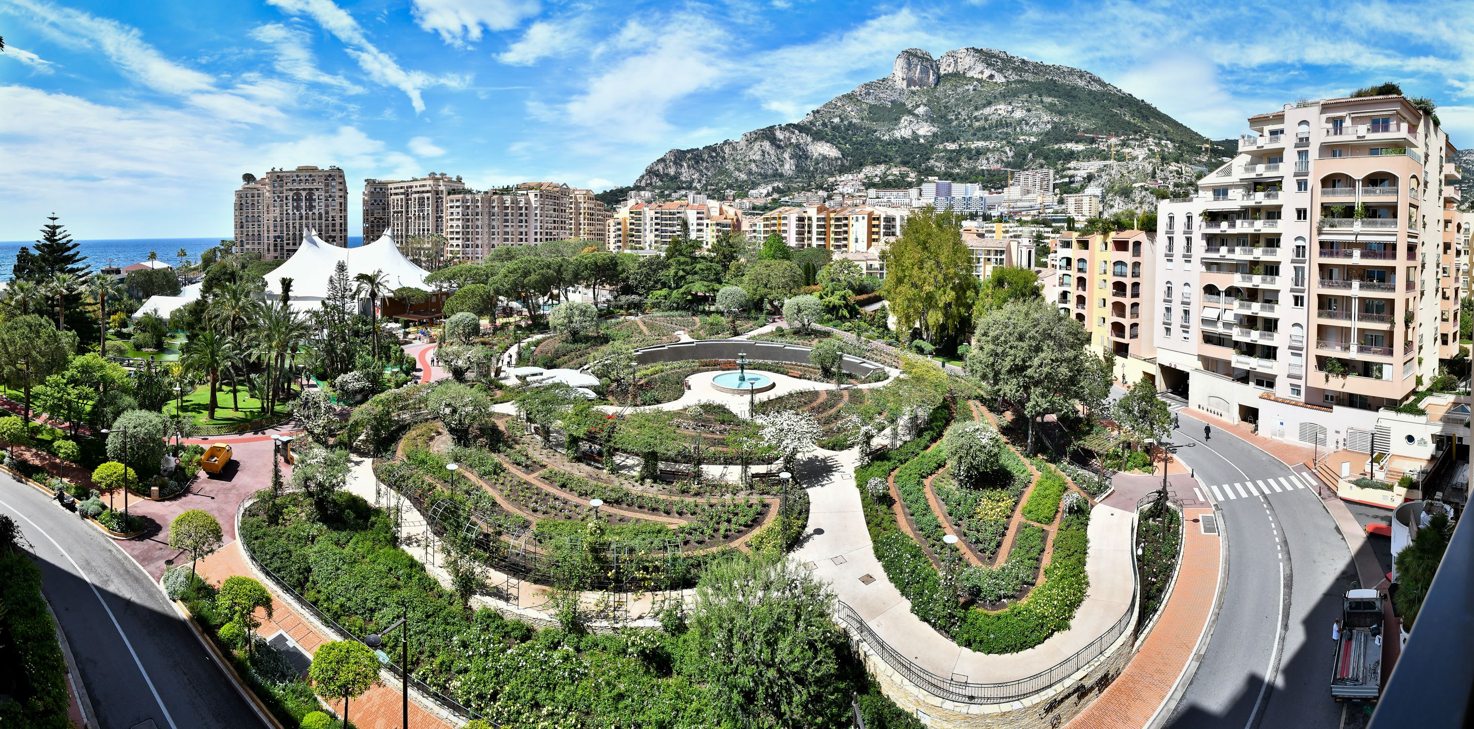 A landscaped garden with a central fountain, surrounded by residential buildings and a mountain in the background.