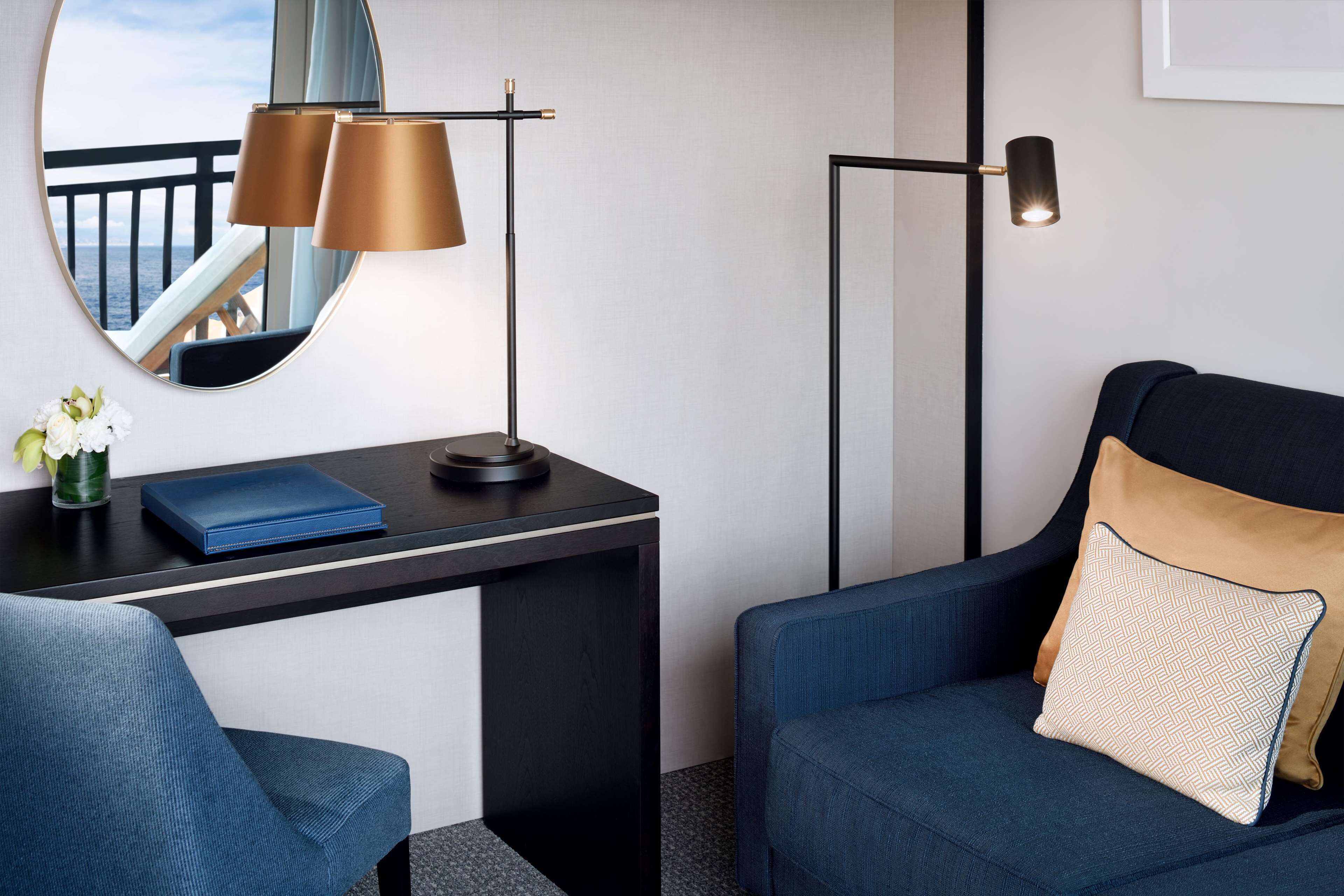 A hotel room with a desk, two chairs, a lamp, a mirror, and a view of the ocean.