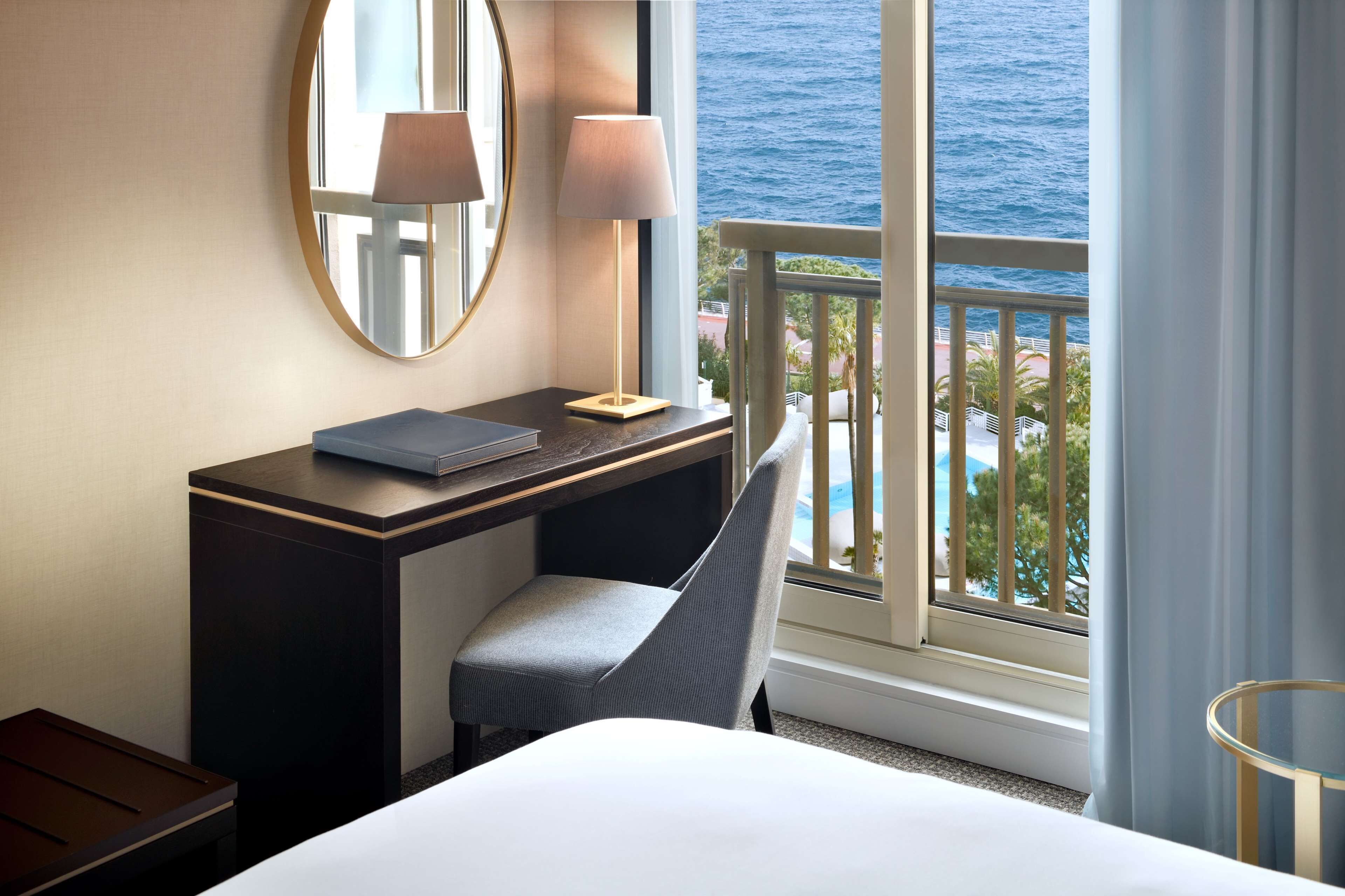 A hotel room with a bed, a desk with a lamp, a chair, a round mirror, and a view of the ocean through a balcony.